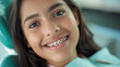 © Elena - A close up photo portrait of smiling teenager girl in dental braces, sitting in dentist’s chair, orthodontist visit, beautiful teeth in brace system, young hispanic woman with blue dentist napkin