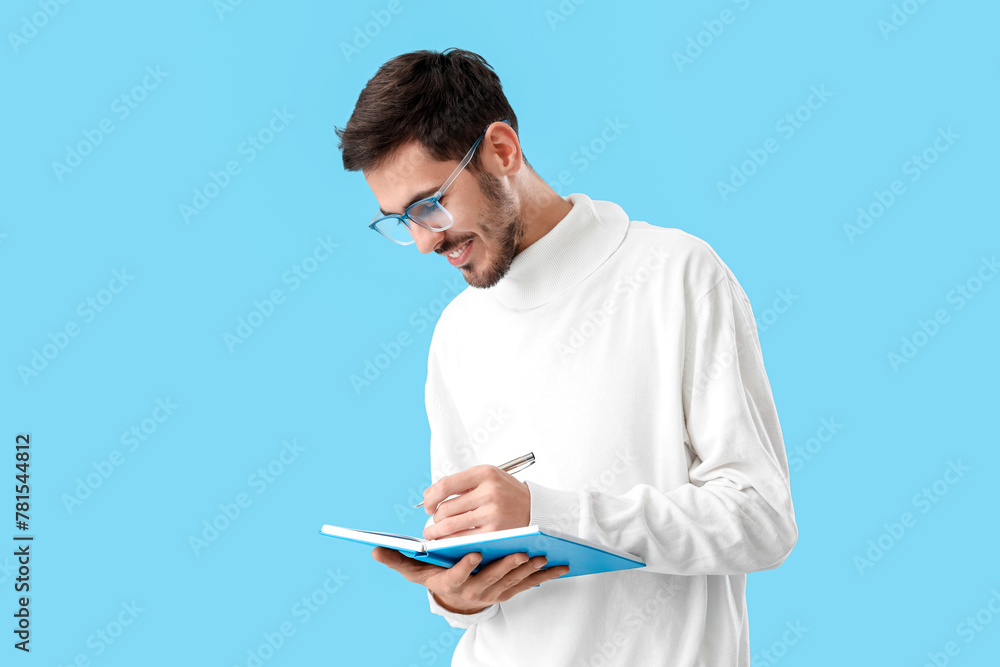 Young man with eyeglasses writing in notebook on blue background