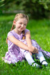 blondes glückliches Mädchen in einem lila Kleid sitz auf dem grünen Gras, schaut in die Kamera, sonniger tag