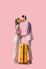 Canvas Print - Beautiful young happy couple of tourists in headphones with suitcase kissing on pink background