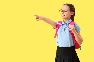 Wall Mural - Happy little schoolgirl with backpack pointing at something on yellow background