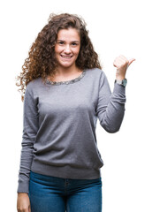 Poster - Beautiful brunette curly hair young girl wearing a sweater over isolated background smiling with happy face looking and pointing to the side with thumb up.