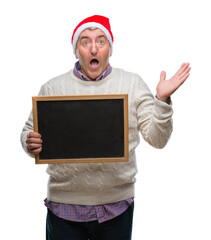 Wall Mural - Handsome senior man wearing christmas hat and holding blackboard over isolated background very happy and excited, winner expression celebrating victory screaming with big smile and raised hands