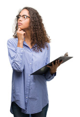 Sticker - Young hispanic business woman holding clipboard serious face thinking about question, very confused idea