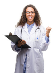 Wall Mural - Young hispanic doctor woman holding a clipboard screaming proud and celebrating victory and success very excited, cheering emotion