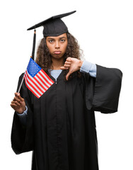 Sticker - Young hispanic woman wearing graduation uniform holding flag of United States with angry face, negative sign showing dislike with thumbs down, rejection concept