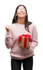 Poster - Young hispanic woman holding a gift screaming proud and celebrating victory and success very excited, cheering emotion