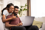 Fototapeta Zachód słońca - Joyful African American couple using a credit card to shop online, sitting comfortably on the couch at home.