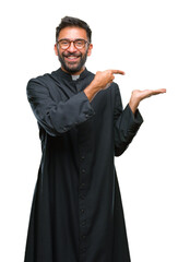 Canvas Print - Adult hispanic catholic priest man over isolated background amazed and smiling to the camera while presenting with hand and pointing with finger.