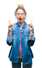 Wall Mural - Young beautiful blonde woman wearing glasses over isolated background amazed and surprised looking up and pointing with fingers and raised arms.