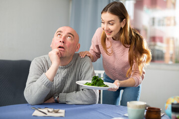 Wall Mural - Portrait of a disgruntled european man sitting at a table in a room, to whom a young female companion brought a plate ..of spinach