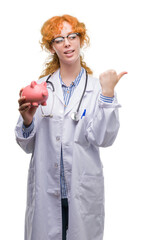 Poster - Young redhead doctor woman holding piggy bank pointing and showing with thumb up to the side with happy face smiling