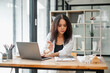 © Satori Studio - Real estate agent examines property documents while working on a laptop in a modern office, with miniature house models on the table.