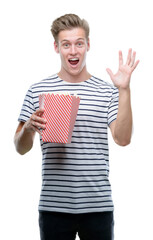 Sticker - Young handsome blond man eating popcorn very happy and excited, winner expression celebrating victory screaming with big smile and raised hands