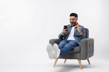 Happy man using smartphone in armchair on white background, space for text