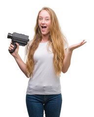 Poster - Blonde woman teenager filming holding super 8 video camera very happy and excited, winner expression celebrating victory screaming with big smile and raised hands