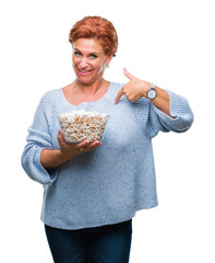 Wall Mural - Atrractive senior caucasian redhead woman eating popcorn over isolated background with surprise face pointing finger to himself