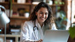 © NooPaew - A cheerful Latino female doctor in a white coat smiles as she works on her laptop in a modern office setting.
