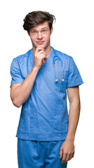Wall Mural - Young doctor wearing medical uniform over isolated background with hand on chin thinking about question, pensive expression. Smiling with thoughtful face. Doubt concept.