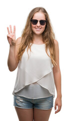 Sticker - Young blonde woman wearing sunglasses showing and pointing up with fingers number three while smiling confident and happy.