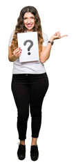 Wall Mural - Young adult woman holding paper with question mark very happy pointing with hand and finger to the side