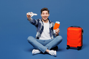 Wall Mural - Traveler man wears casual clothes sit near bag hold passport ticket air plane mockup isolated on plain blue background. Tourist travel abroad in free time rest getaway Air flight trip journey concept