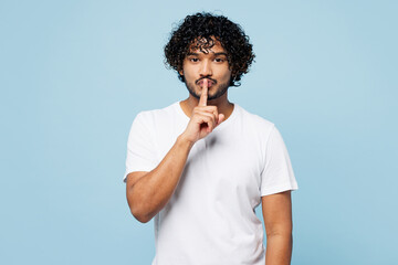 Wall Mural - Young secret Indian man he wear white t-shirt casual clothes say hush be quiet with finger on lips shhh gesture isolated on plain pastel light blue cyan background studio portrait. Lifestyle concept.
