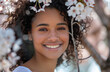 A close-up portrait of an attractive young woman with curly hair, smiling and looking at the camera while standing in front of blooming cherry blossoms on a sunny day.