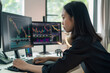 © Kamonwan - An Asian woman is captured at her home office setup, deeply focused on stock or cryptocurrency trading on her computer.