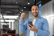 © Tetiana - Portrait of a happy Muslim man standing in a modern office, holding a phone in his hand and rejoicing in success and news, showing a hand gesture, looking at the camera