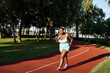 A curvy African American woman in sportswear runs energetically on a track in a green park.
