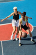 © LIGHTFIELD STUDIOS - Two young women, friends and athletes, engaged in a competitive game of basketball on an outdoor court in the summer.