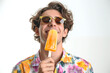 © Concept Island - Young man with sunglasses and flowered summer shirt eating and enjoying an orange popsicle, isolated on a white background.