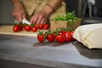 Selective focus on a bunch of red ripe tomato cherry on the kitchen table against the background of a male chef chopping vegetable, cooking salad for dinner at home. Copy advertising space