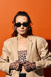 A fashionable woman in sunglasses poses in a tan jacket and leopard print gloves against an orange studio backdrop.