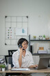 © apichat - Cheerful businesswoman in white shirt using smartphone with laptop and documents on her office desk.