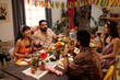 Group of young friends having chat by served festive table while enjoying dinner with homemade food in living room decorated with pennants