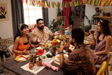 Fototapeta  - Group of young friends having chat by served festive table while enjoying dinner with homemade food in living room decorated with pennants