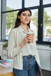 © LIGHTFIELD STUDIOS - A modern businesswoman stands by a window, holding a cup of coffee.