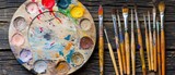 Fototapeta Mapy -   A tight shot of a palette bearing paint, adjacent to an array of paintbrushes on a rustic table against a wooden backdrop