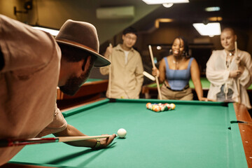 Wall Mural - Over shoulder view of African American man playing pool and hitting ball with cue stick with diverse group of people in background copy space
