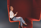 Fototapeta Na drzwi - Cheerful senior businesswoman using a smartphone, sitting on a modern red bench with documents, enjoying a light moment in a creative office space.