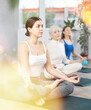 Sedulous women doing lotus pose of yoga on black mat in light gym room with pot plants