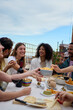 © CarlosBarquero - Vertical. Group of multi-ethnic young friends enjoying meal gathered on rooftop. Happy millennial people having fun drinking red wine and eating celebrating a party event on summer day. Copy space