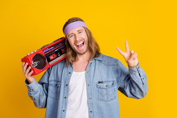 Wall Mural - Portrait of cool young man hold boombox show v-sign wear denim shirt isolated on yellow color background