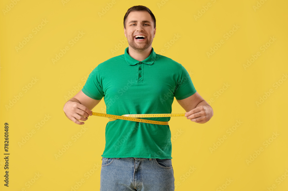 Young man measuring his waist on yellow background. Weight loss concept