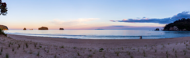 Wall Mural - A Breathtaking View of Whangamata Beach as the Sun Sets, Casting a Spectacular Purple Glow Across the Sky, with People Walking Along the Shoreline