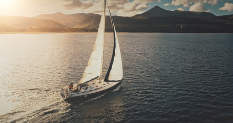 Closeup aerial sailboat cruise at sun sea bay. Sunshine reflection at water near mountains ocean coast of Arran island, Scotland, Europe. Luxury yachts with passengers. Summer nature seascape