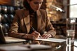 A businesswoman in an office, sitting at her desk and writing on paper with a pen, wearing a brown blazer over a black turtleneck sweater, a brick wall behind the woman, a computer screen