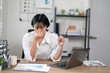 © Wasana - Exhausted female office worker feeling stressed, holding glasses, with laptop and documents on desk.